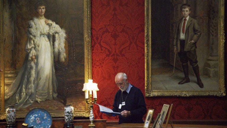 A visitor is observing some notes in a folder. He's stood in front of two grand portraits in Cragside's Drawing Room.